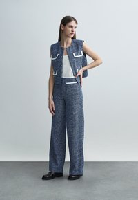 Femeie în picioare cu mâna pe șold, purtând o vestă albastră din țesătură tweed fără mâneci și pantaloni largi asortați, cu pantofi negri.