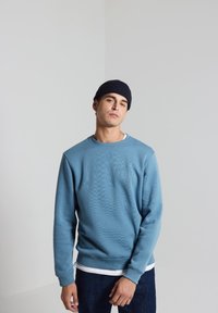 Blauwe sweatshirt van gestructureerde stof, ronde halslijn, ribgebreide manchetten en zoom. Draagt over een wit T-shirt en gecombineerd met donkere jeans.