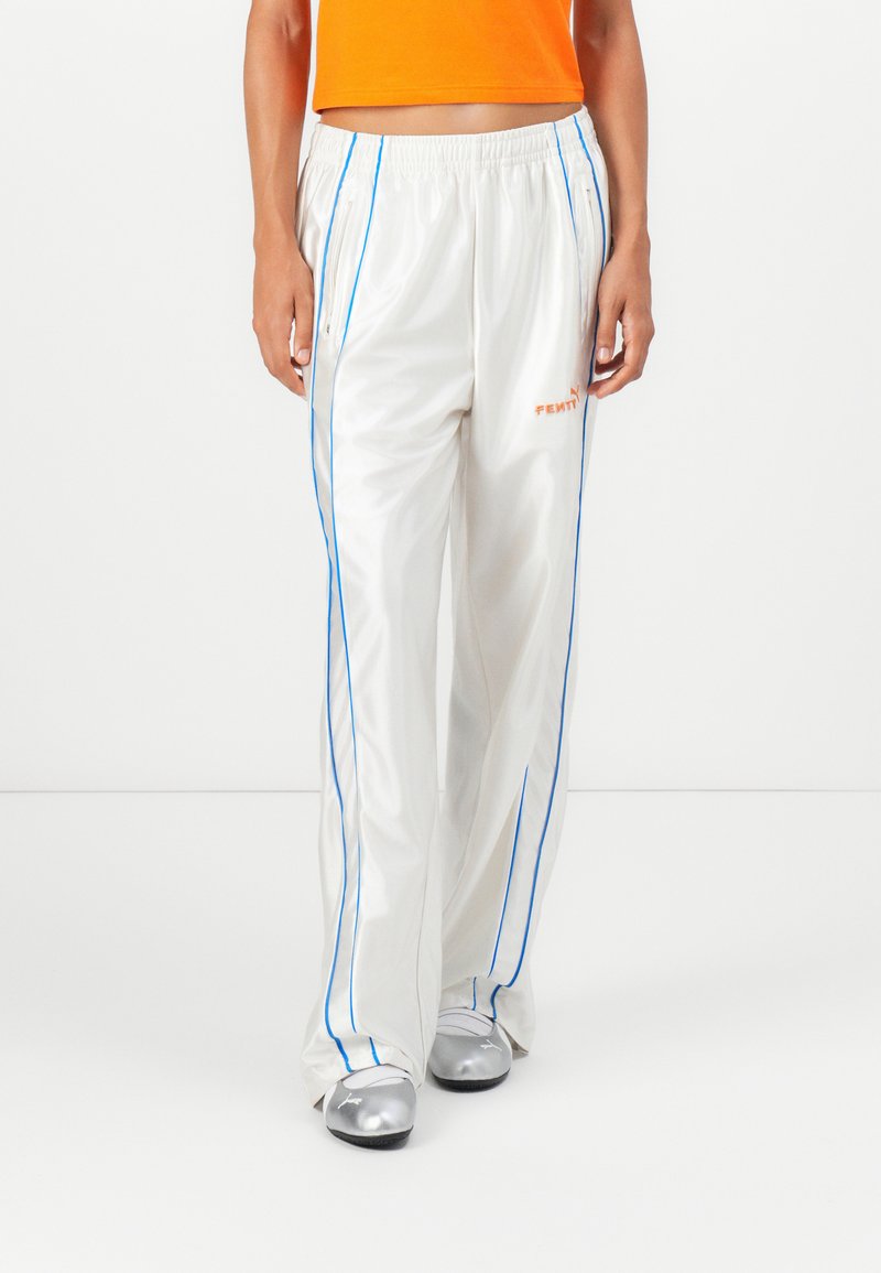 Persoon in witte satijnen trainingsbroek met blauwe piping, oranje crop top en zilveren Puma-schoenen, staand tegen een witte achtergrond.