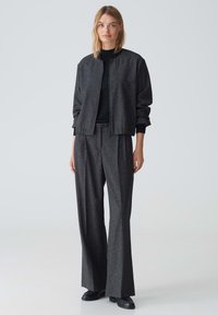 Grau strukturierte Bomberjacke, kombiniert mit hoch taillierten, weit geschnittenen Hosen, beide aus einem ähnlichen Stoff, übereinander getragen mit einem schwarzen Oberteil.