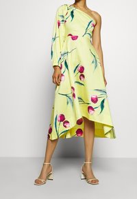 Žena v žlutých midi šatech na jedno rameno s růžovým květinovým vzorem a dlouhým nařaseným rukávem, doplněná o bílé sandály na podpatku.