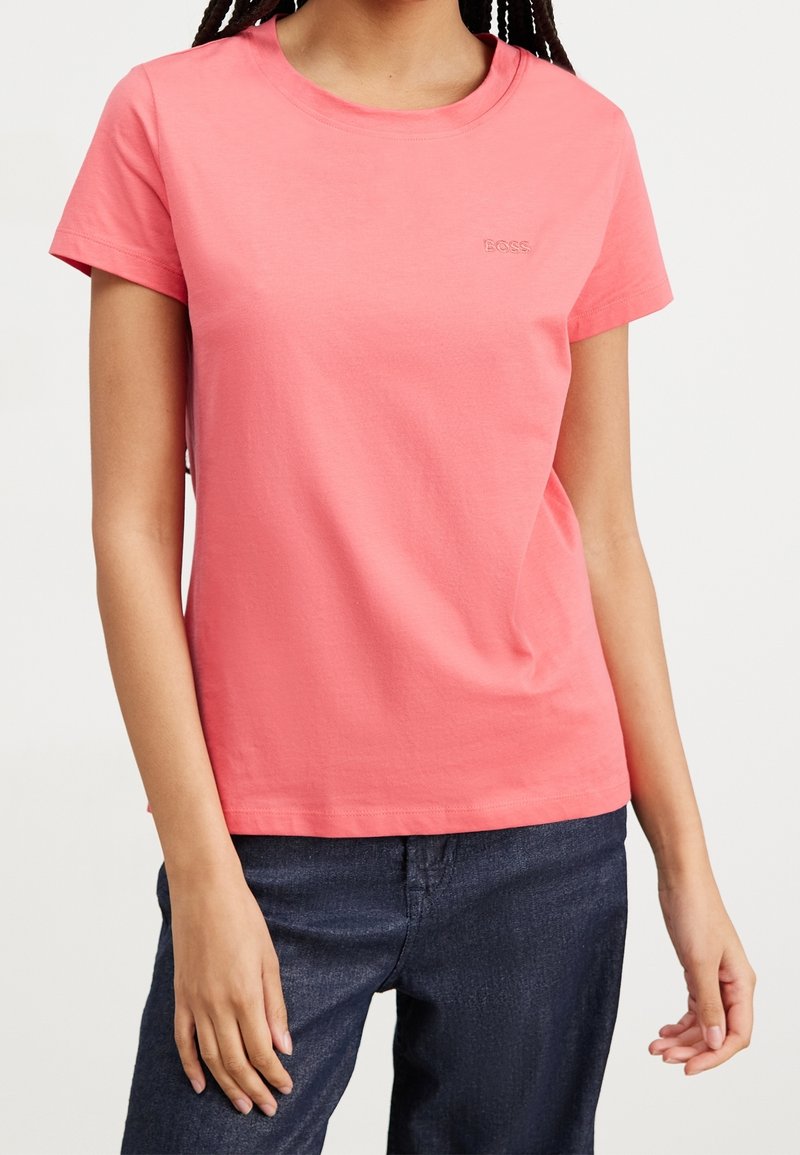 Kvinna som bär en korallrosa kortärmad skjorta med liten präglad "BOSS"-logotyp, tillsammans med mörkblå jeans, stående mot vit bakgrund.