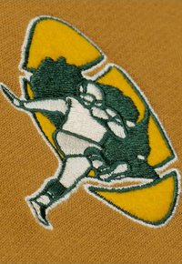 Besticktes Abzeichen mit einer Figur in Grün und Weiß, schwarz umrandet, vor einem gelben Hintergrund auf einem strukturierten braunen Stoff.
