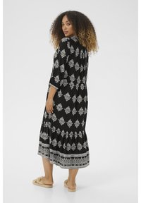 Schwarzes Maxikleid mit weißem Blumenmuster, fließendem Design, drei Viertelärmeln und gerafftem Saum. Getragen mit hellen, geknoteten Sandalen.