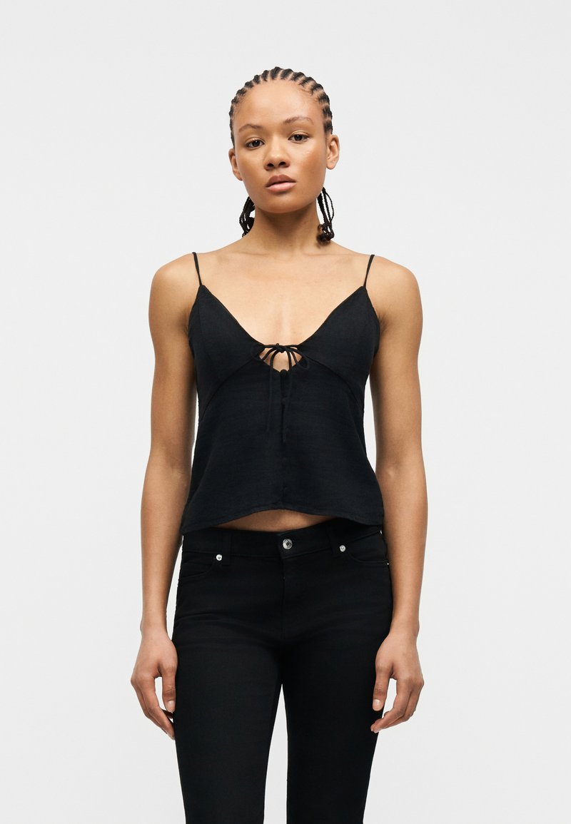 Γυναίκα με πλεξούδες cornrow, φορώντας μαύρο crop top χωρίς μανίκια με λεπτές τιράντες και μαύρο στενό παντελόνι, στέκεται μπροστά σε ένα απλό φωτεινό φόντο.