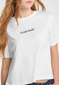 Camiseta de algodón blanca con mangas cortas, que presenta un logo negro de "GUESS JEANS" impreso en el pecho. Corte recto en el dobladillo.