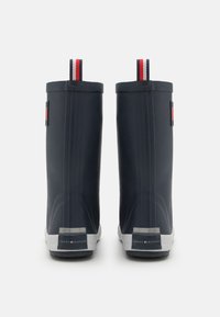 Bottes de pluie en caoutchouc bleu marine avec des semelles blanches, des rainures verticales et une languette de tirage accentuée en rouge. Présente un logo à l'arrière. Texture lisse.