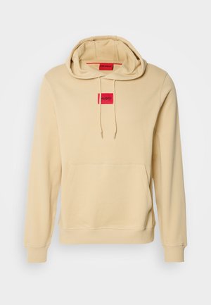 Beige hoodie van zacht materiaal, met een kangoeroezak, ribgebreide boorden en een rood rechthoekig logopatch op de borst.