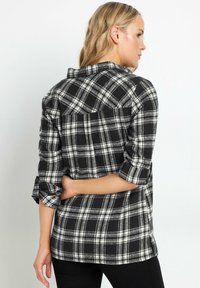 Camisa de franela a cuadros en blanco y negro con mangas enrolladas, que presenta un yugo en la parte posterior y aberturas laterales inferiores para mayor comodidad.