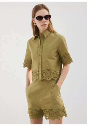 Vrouw draagt een olijfgroen overhemd met korte mouwen en knopen, en bijpassende korte broek met ronde uitsnijdingen, zwarte zonnebril en gouden creolen.