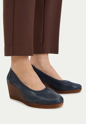 Kobieta w brązowych spodniach do połowy łydki i granatowych koturnach z teksturowanymi drewnianymi podeszwami, stojąca na jasnym tle.