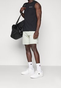 Top sin mangas negro, pantalones cortos verdes claros, zapatillas blancas, calcetines blancos con el logo de Champion, llevando una bolsa de gimnasio negra.