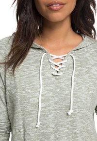 Hoodie gris clair texturé avec un col à lacets et des cordons blancs. Présente une coupe décontractée et une capuche.