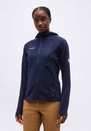 Žena v tmavomodrej Mammut mikine na zips s otvorom na palce a béžových nohaviciach, stojaca proti jednoliatemu bielemu pozadiu.