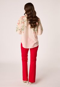 Lichtroze blouse met bloemenborduursel in groen en rood, gecombineerd met strakke rode broeken. Kenmerken zijn een gebogen zoom en lange mouwen.