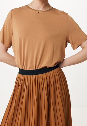 Vrouw draagt een bruin topje met korte mouwen, ingestopt in een bruin gerimpelde rok met hoge taille en een zwarte tailleband, handen op de heupen.