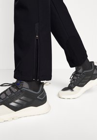 Czarne sportowe spodnie z zamkami błyskawicznymi i nakładkami na kolanach, w połączeniu z czarno-białymi trampkami z elementami z siateczki i gumy oraz teksturowanymi sznurowadłami.