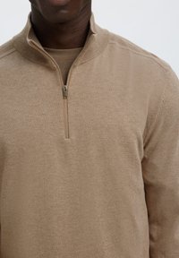 Mann trägt einen beigen Halb-zip-Pullover über einem passenden beigefarbenen Hemd, sichtbar vom Hals bis zur Mitte der Brust vor einem einfarbigen Hintergrund.