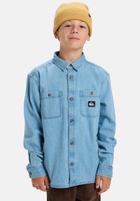 Niño con una camisa de mezclilla azul claro de botones y bolsillos en el pecho, usando un gorro de lana amarillo, de pie contra un fondo sencillo.