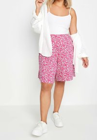 Růžové květinové šortky s bílými květinovými vzory v kombinaci s bílým tílkem a oversized bílou košili. Celý vzhled dokončují bílé tenisky.