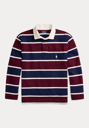 Femme vêtue d’un maillot de rugby rayé, un pull gris drapé sur les épaules, un pantalon gris plissé, une ceinture marron, des chaussures beige, des lunettes de soleil, tenant un sac à main marron.
