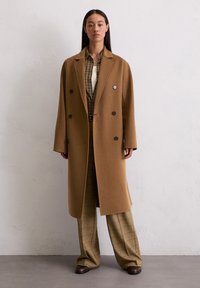 Femme portant un long manteau couleur sable sur une chemise à carreaux marron et un pantalon à carreaux large, se tenant contre un mur blanc texturé.