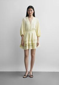 Kvinne med langt svart hår iført en blekgul, lagdelt kjole med 3/4-ermer og svarte, stroppelignende heeled sandaler, stående på et grått gulv.