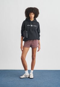 Fiatal nő áll, fekete Nike Shox kapucnis pulóvert, bordó Nike rövidnadrágot, fehér zoknit, fekete talpú fehér sportcipőt és egy kis fekete oldaltáskát viselve.