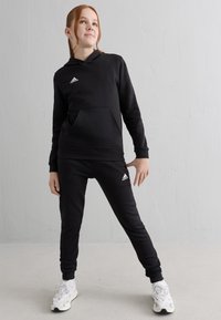 Jeune personne portant un hoodie Adidas noir et un pantalon de survêtement, avec des baskets blanches, se tenant devant un mur clair, regardant vers le haut.