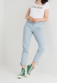 Jeans de tiro alto en azul claro con detalles desgastados en los tobillos, combinados con una camisa blanca de manga corta con un escrito dorado y zapatillas verdes.