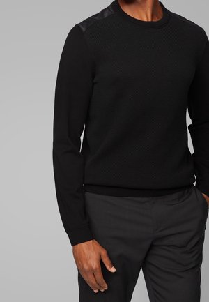 Mężczyzna ubrany w czarny teksturowany sweter z długim rękawem z błyszczącymi naszywkami na ramionach oraz ciemne spodnie na jednolitym szarym tle.