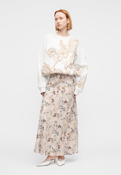 Ανοιχτό γκρι φαρδιά φούτερ με χρυσαφένιες κεντητές ανθισμένες λεπτομέρειες, συνδυασμένο με αέρινη, εμπριμέ μακρυά φούστα σε απαλά παστέλ χρώματα και λευκά ψηλοτάκουνα παπούτσια.