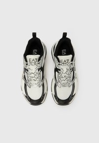 Zapatillas deportivas con parte superior de malla en blanco y negro. Presentan detalles plateados y un diseño de cordones con patrón. Suela de goma texturizada.