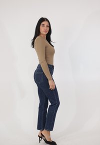 Top beige de manga larga, jeans de talle alto en azul oscuro y tacones negros; fondo liso; cuerpo en vista de perfil, mostrando ajuste y silueta.