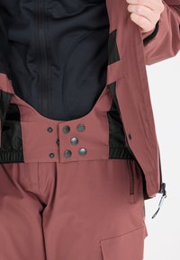 Die kastanienbraune Außenjacke hat ein schwarz gefüttertes Innenfutter, einen elastischen Bund und einen Druckknopfverschluss mit vier schwarzen Knöpfen. Seitliche Reißverschlusstasche.