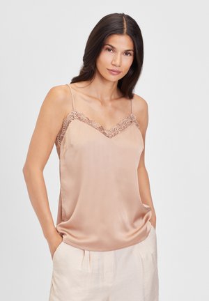 Satin cami i lys beige med blondekant ved halsudskæringen. Har tynde stropper og en afslappet pasform. Modellen stiller sig med hænderne i lommerne.