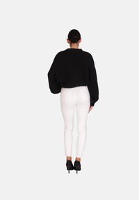 Sort oversized strikket sweater med sænkede skuldre, kombineret med tætsiddende hvide jeans. Modellen har sorte højhælede sko på og ses bagfra.