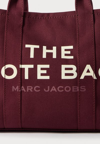 Bordowa torba na ramię z płótna z białym napisem "TORBA" oraz "MARC JACOBS", z mocnymi uchwytami i teksturowanym wykończeniem.