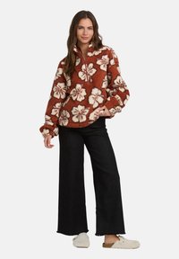 Pullover in pile terracotta con motivi floreali bianchi, colletto alto e chiusura con zip. Abbinato a jeans neri a gamba larga e pantofole pelose.