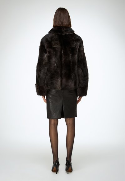 Veste en fausse fourrure marron foncé avec un large col. Associée à une jupe en cuir noire ajustée et des chaussures à talons noirs. Texture lisse.