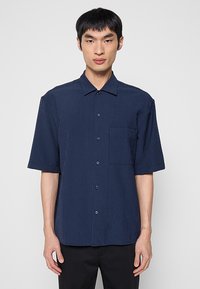 Camisa azul marino de manga corta con rayas verticales, cierre frontal con botones, cuello y un bolsillo en el pecho. La tela parece ligera y transpirable.