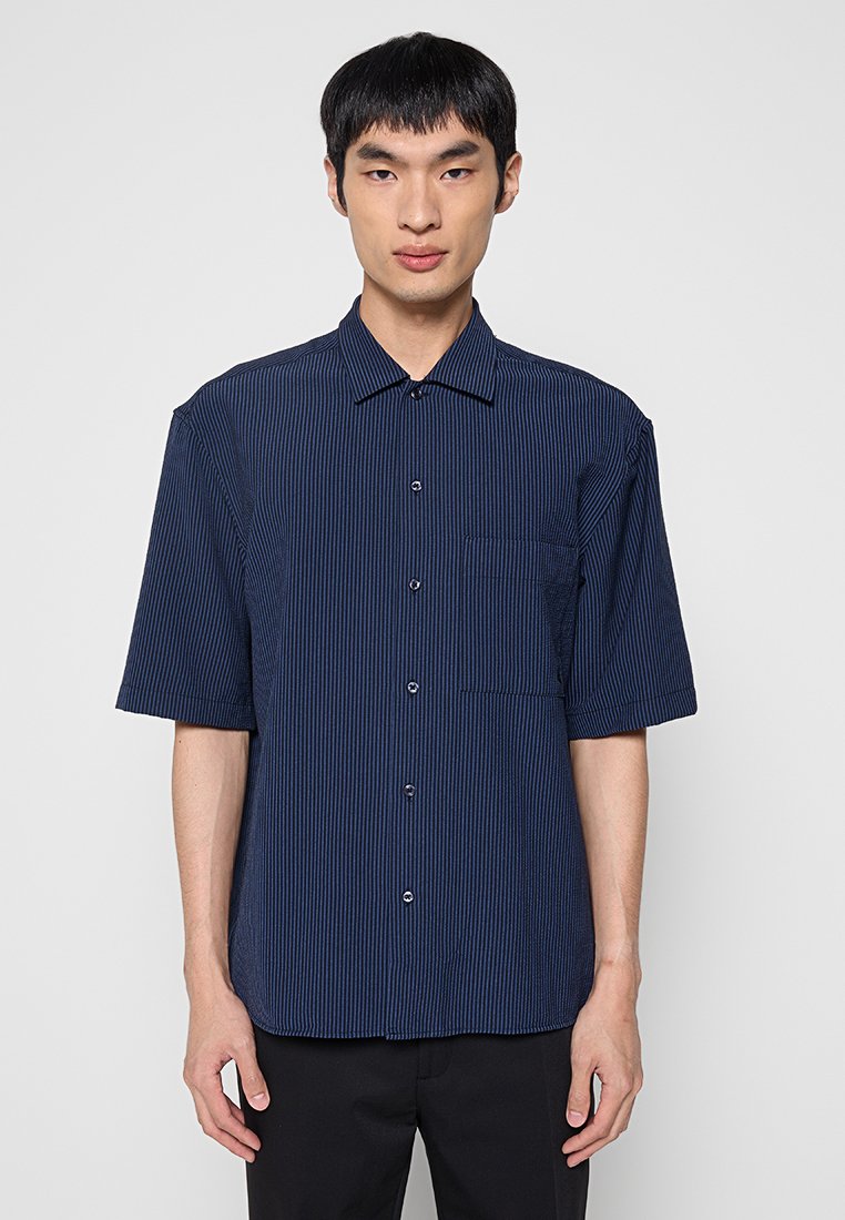 Camisa azul marino de manga corta con rayas verticales, cierre frontal con botones, cuello y un bolsillo en el pecho. La tela parece ligera y transpirable.