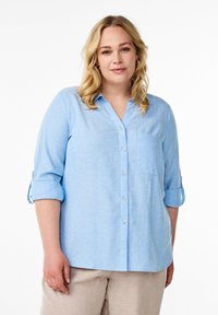 Camicia button-up in lino azzurro chiaro con scollatura a V, maniche lunghe arrotolate e taschino sul petto. Tessuto morbido e vestibilità comoda.