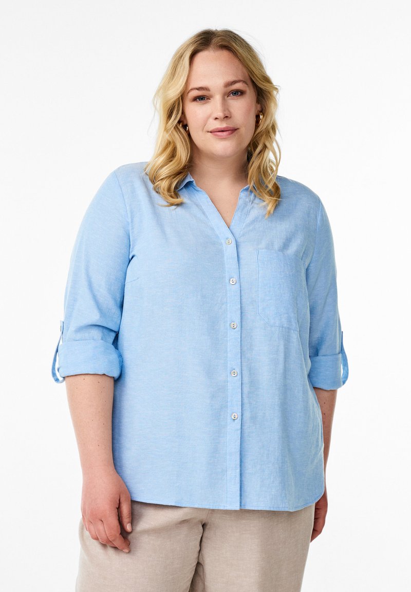 Camicia button-up in lino azzurro chiaro con scollatura a V, maniche lunghe arrotolate e taschino sul petto. Tessuto morbido e vestibilità comoda.