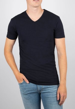 Man in een eenvoudig zwart V-hals T-shirt met korte mouwen en lichtblauwe jeans, met één hand in zijn zak, tegen een witte achtergrond.