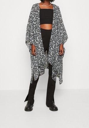 Osoba nosząca czarny krótki top, czarne spodnie z rozcięciami z przodu, czarne buty i luźny kardigan w czarno-biały kwiatowy wzór.