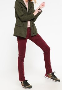 Osoba ubrana w oliwkową zieloną kurtkę, jasnoróżową koszulę, bordowe spodnie i zielone trampki z czarnymi gwiazdkami, stojąca z nogami rozstawionymi.