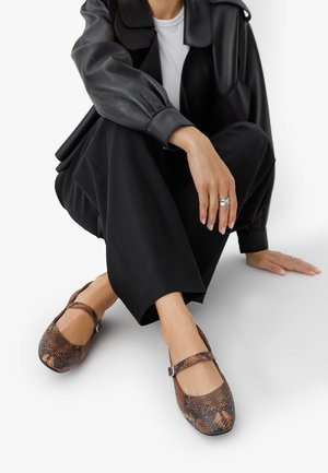 Person wearing black wide pants, black leather jacket, and brown snakeskin-patterned Mary Jane shoes, sitting on the floor with one hand resting.
