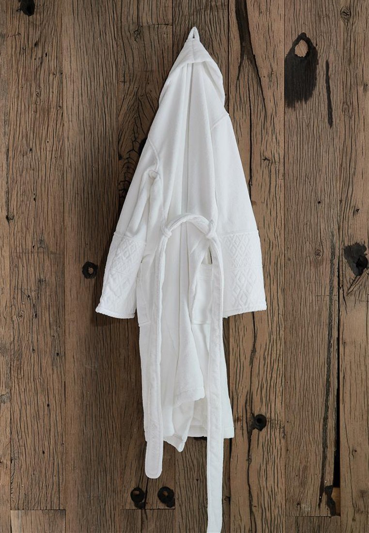 Accappatoio bianco in cotone con cappuccio, cintura in vita e dettagli testurizzati sulle maniche, appeso contro un muro di legno rustico.