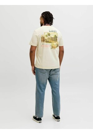 Bărbat cu dreadlocks purtând un tricou bej cu grafică, blugi albaștri, adidași negri, stând cu fața spre spate pe un fundal simplu.